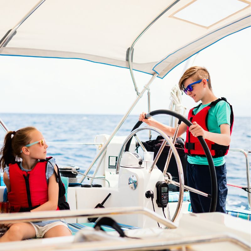 Two children wearing life jackets on a sailboat; one boy steering the wheel while a girl sits nearby, both enjoying a sunny day at sea.